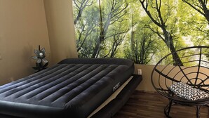 A room with a bed, a chair, a ceiling fan, and a large window with a forest view.