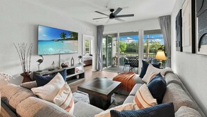 Ein geräumiges Wohnzimmer mit einem großen Flachbildfernseher, bequemen Sitzmöglichkeiten, einem Deckenventilator und Blick ins Freie durch große Schiebeglastüren.