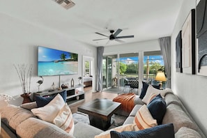 Een ruime woonkamer met een grote flatscreen televisie, comfortabele zitplaatsen, een plafondventilator en uitzicht op de natuur door grote schuifdeuren.