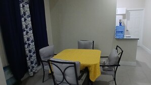 Uma área de refeições com mesa coberta por um tecido amarelo, duas cadeiras, um dispensador de água e uma porta.