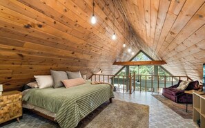 Una camera da letto con soffitto in legno, un letto con cuscini, un comodino e una finestra vista su del verde.