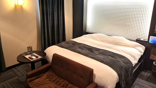 Ein Hotelzimmer mit einem großen Bett, einem Kopfteil mit geometrischem Muster, einem kleinen Nachttisch mit Fernbedienung, einem braunen Sessel und einem Fenster mit einem geometrischen Vorhang.