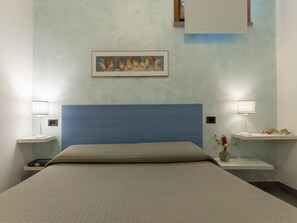 Habitación de hotel con cama, dos mesitas de noche con lámparas, un cuadro colgado en la pared y una pequeña estantería con objetos decorativos.