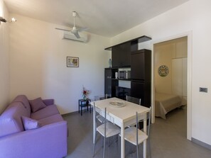 Una sala moderna con un sofá morado, una mesa de comedor con sillas, un ventilador de techo y una zona de cocina con armarios y electrodomésticos.
