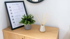 Un letrero de normas de la casa enmarcado, una planta en maceta y un difusor de aroma de caña sobre un mueble de madera.