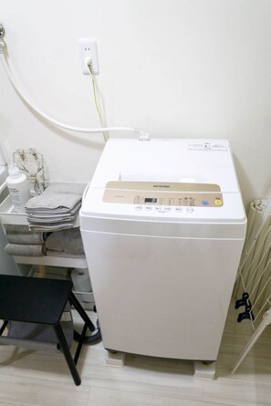Eine kompakte, weiße Waschmaschine mit Bedienfeld, ein kleiner Tisch mit gefalteten Handtüchern und ein Stuhl.