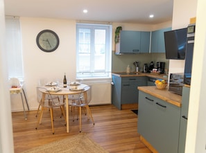 Une cuisine avec une table à manger, des chaises, une horloge et une fenêtre avec des stores.