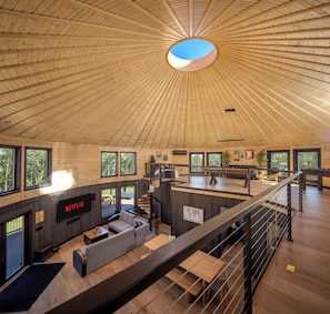 Un interior espacioso de madera con techo alto, una escalera, una sala de estar con sofá y mesa de centro, y un área de cocina.