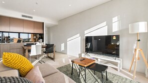Un salon moderne doté d’un grand téléviseur à écran plat, d’un canapé, d’une table basse et d’un coin repas avec des chaises, et une cuisine en arrière-plan.