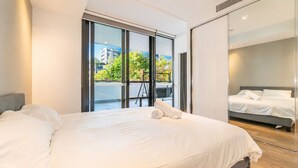 Habitación de hotel moderna con una cama grande, un balcón que ofrece vistas a edificios y árboles, y una puerta corrediza de vidrio.