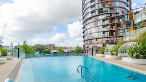 Ein Schwimmbecken mit Glasgeländer, umgeben von Topfpflanzen und einem modernen Gebäude mit einer gewölbten Fassade.