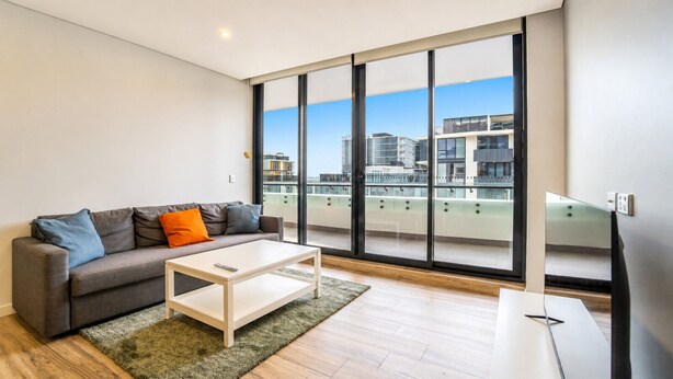 A modern living room with a sofa, coffee table, and large windows offering a city view. - Eastgardens 2BR/2BA | 2 Parking + Shops (Eastgardens)