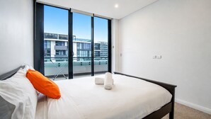 A modern hotel room with a large bed, a view of a building through floor-to-ceiling windows, and a balcony. - Eastgardens 2BR/2BA | 2 Parking + Shops (Eastgardens)