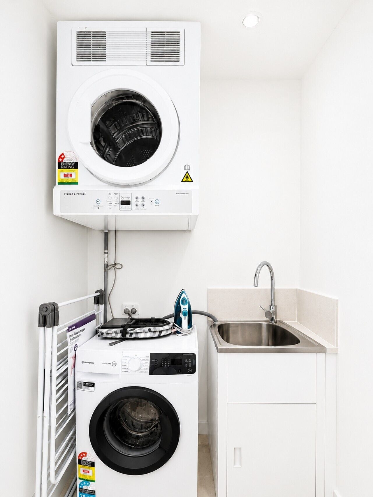 A compact laundry area with a front-loading washer and dryer, a small ironing station, and a sink.