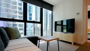 Ein modernes Wohnzimmer mit einer Couch, einem kleinen Tisch, einem Fernseher und großen Fenstern mit Blick auf die Stadt.