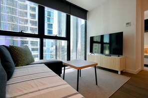 Ein modernes Wohnzimmer mit einer Couch, einem kleinen Tisch, einem Fernseher und großen Fenstern mit Blick auf die Stadt.