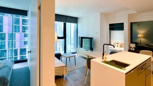 Ein modernes Hotelzimmer mit Bett, Schreibtisch, Fernseher, einer kleinen Küchenzeile und großen Fenstern mit Blick auf die Stadt.