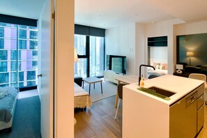 Ein modernes Hotelzimmer mit Bett, Schreibtisch, Fernseher, einer kleinen Küchenzeile und großen Fenstern mit Blick auf die Stadt.