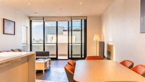 Una habitación de hotel moderna con un gran ventanal corredero que da a un balcón, una mesa de comedor de madera, un sofá y un televisor.