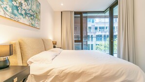 Ein Hotelzimmer mit Bett, Nachttisch, Lampe, einem Gemälde und Blick auf Gebäude durch das Fenster.