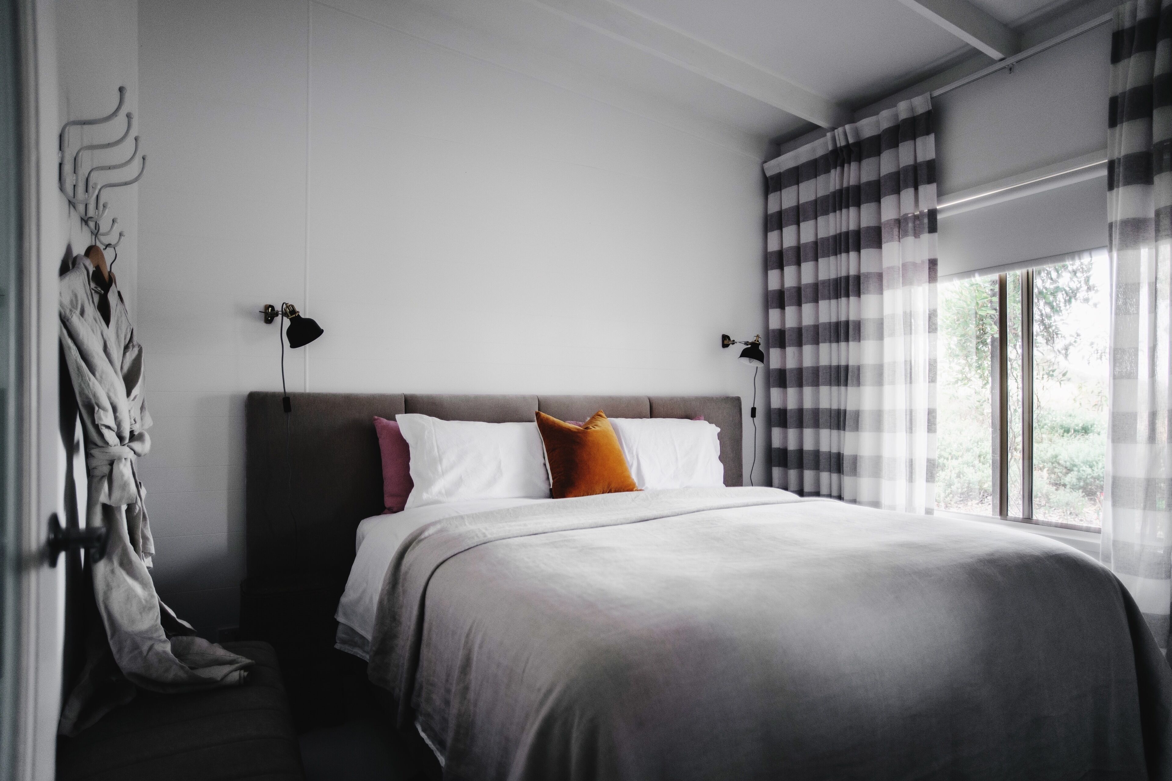 A modern bedroom with a large bed, two bedside lamps, a window with curtains, and a coat rack with a robe.
