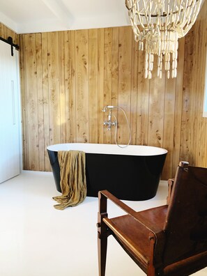 A modern bathroom with a wooden wall, a freestanding bathtub, a chandelier, and a leather chair.