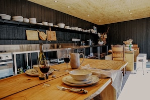 A wooden dining table with plates, a wine glass, and a pitcher in a modern kitchen with dark wooden walls and a wooden ceiling.