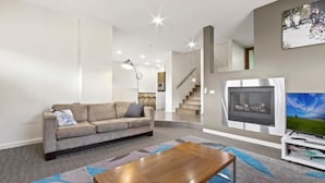 A modern living room with a sofa, a coffee table, a fireplace, a TV, and a staircase leading to another level. - Blue Ice Chalet (Dinner Plain)
