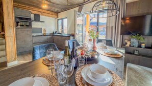 A dining area with a wooden table set for a meal, a kitchen in the background, and a large window with a view.