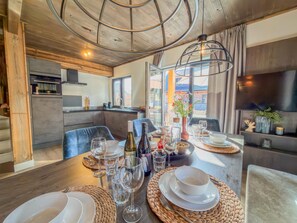 A dining area with a wooden table set for a meal, a kitchen in the background, and a large window with a view. - Spacious chalet by an alpine lake in Carinthia (Afritz am See)