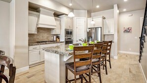 Cocina moderna con armarios blancos, encimeras de granito, electrodomésticos de acero inoxidable y una isla central con taburetes de bar.