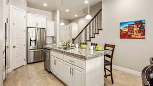 Una cocina con gabinetes blancos, refrigerador de acero inoxidable, encimeras de granito, lavavajillas integrado y un comedor con sillas de madera.