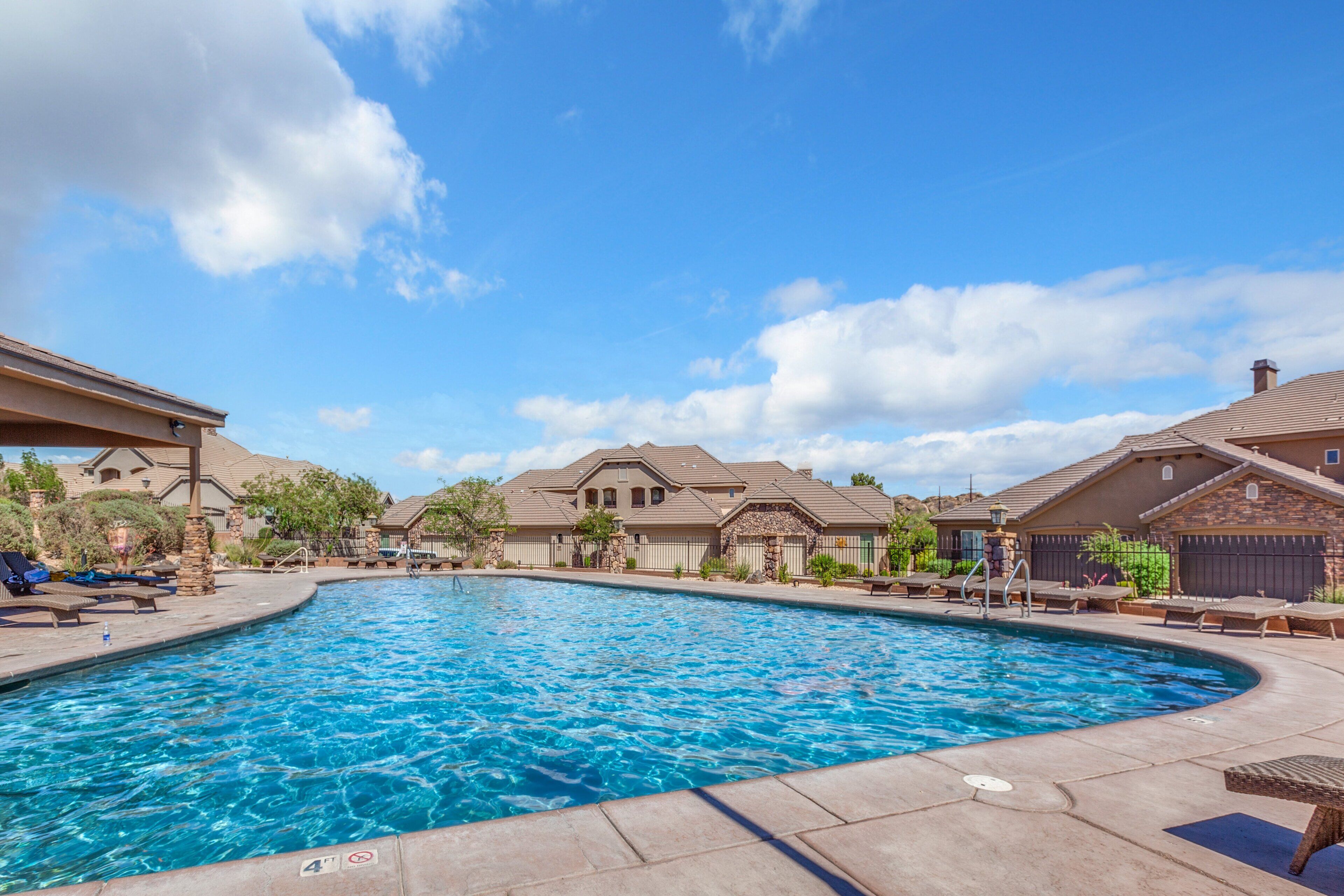A swimming pool with lounge chairs and a covered area, surrounded by houses with a clear sky above.
