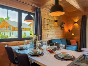 Ein Holzinnenraum mit einem für vier Personen gedeckten Esstisch, einem blauen Sofa und Blick auf ein Haus durch große Fenster.