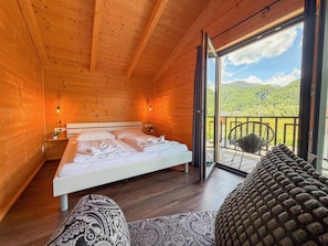 Ein Schlafzimmer einer Holzhütte mit Bett, Nachttischen, einer Couch und einem Balkon mit Blick auf grüne Hügel.