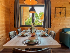 Una habitación con paneles de madera, una mesa de comedor lista para seis comensales, un sofá y un amplio ventanal con cortinas.