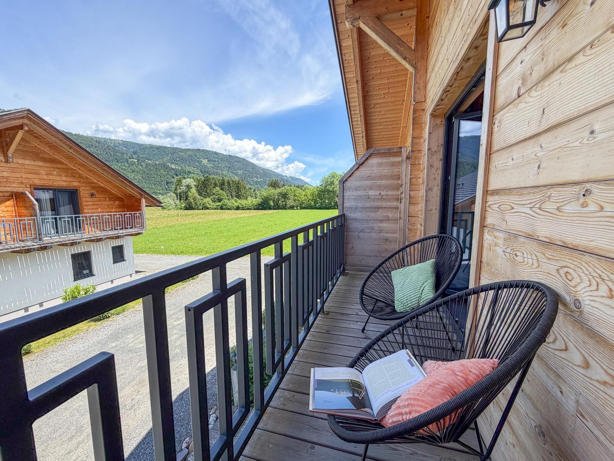 A wooden balcony with a view of a green field and mountains, a wicker chair with a cushion, and a wooden building with a balcony.