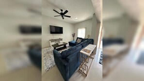 Een woonkamer met een modulaire bank, een televisie, een plafondventilator en een eethoek met een tafel en stoelen.