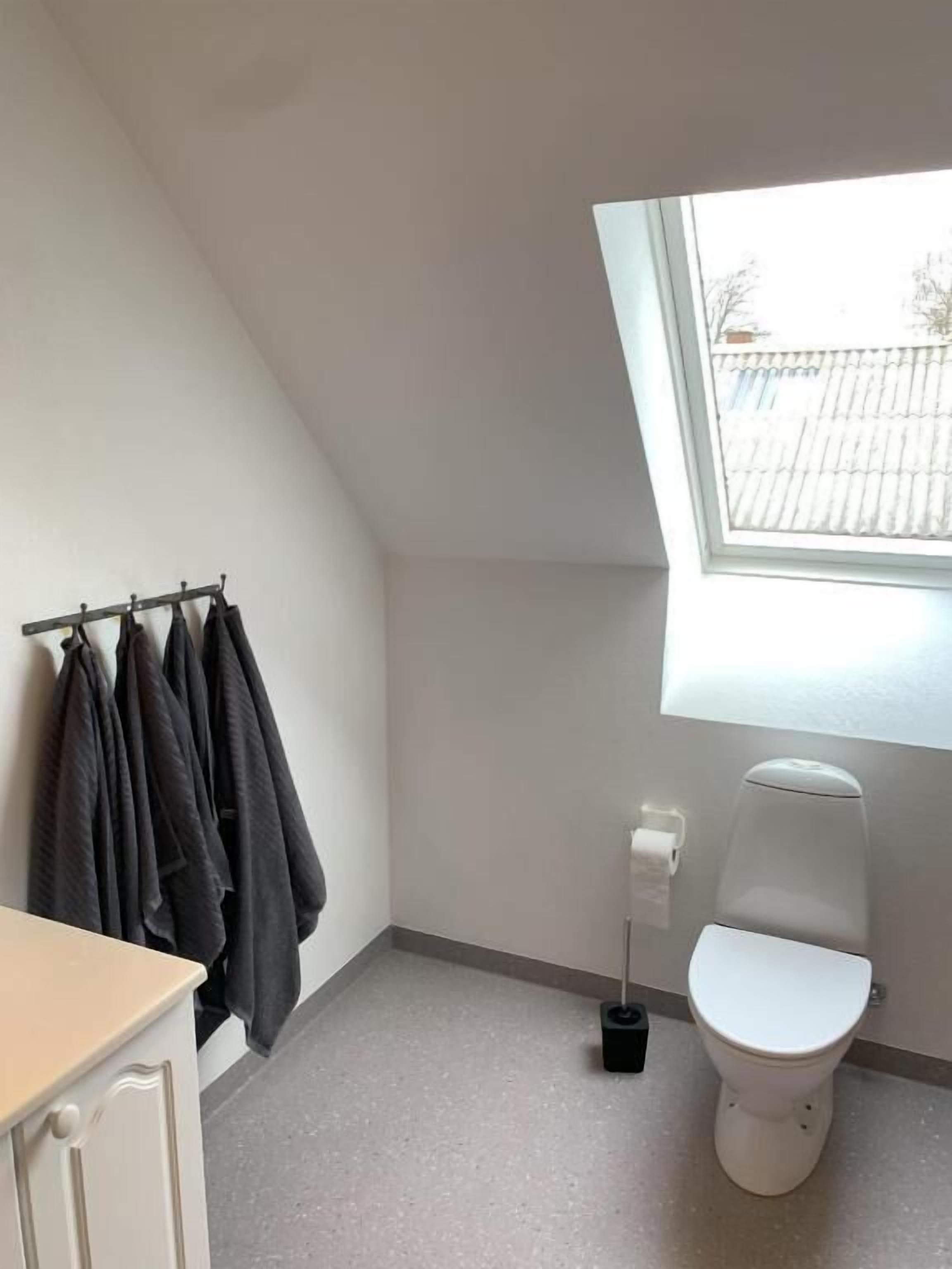 Ein kleiner, heller Raum mit schrägverlaufender Decke, einer Toilette, einem Fenster und einem Kleiderhaken mit mehreren Mänteln.