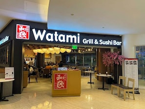 A restaurant named 'Watami Grill & Sushi Bar' with a signboard, a reception desk, and a seating area with tables and chairs.