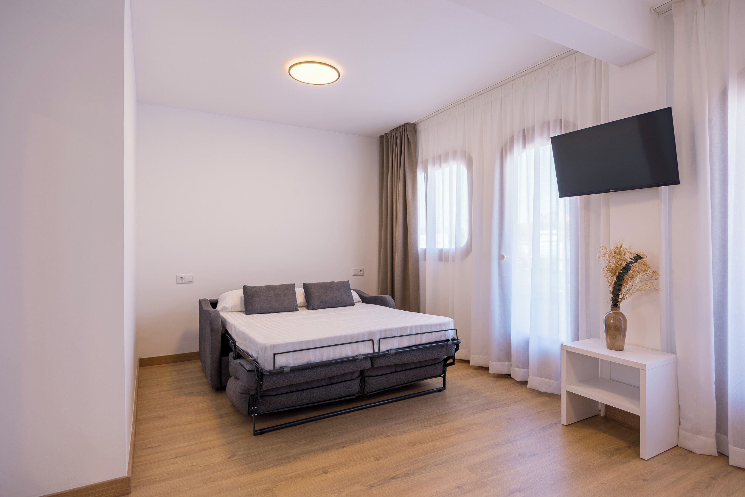 Un dormitorio moderno con cama, televisor, mesita de noche y ventana con cortinas.