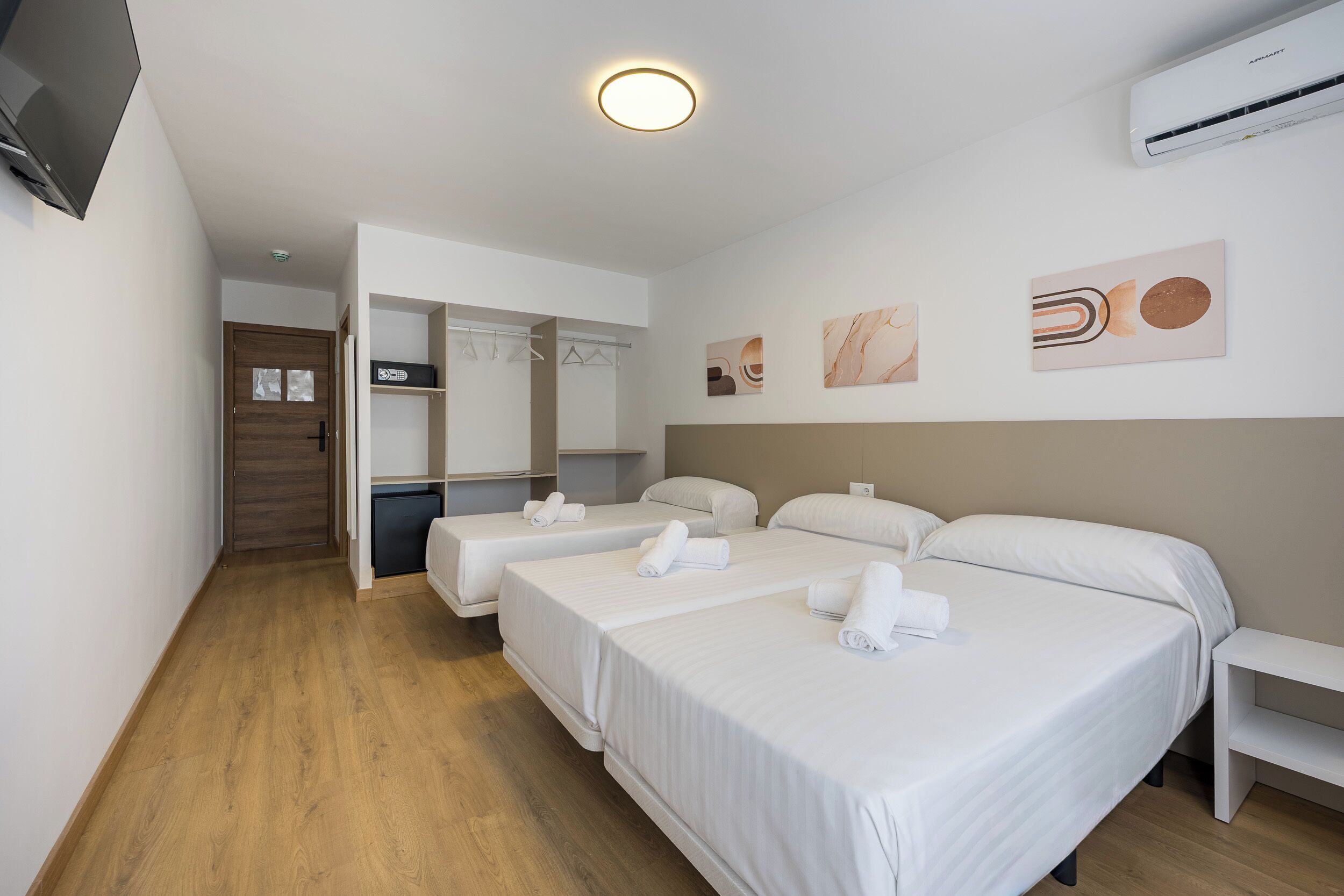 A hotel room with three beds, a wooden floor, a flat-screen TV, a wall-mounted air conditioner, and a hallway leading to a bathroom.