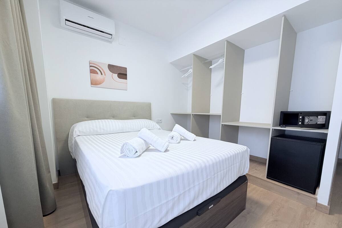 A modern hotel room with a bed, air conditioning unit, artwork, wardrobe, and a small black refrigerator.