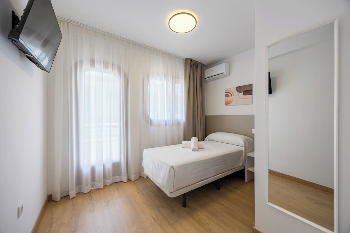 A hotel room with a bed, a TV, a ceiling light, and a window with curtains.