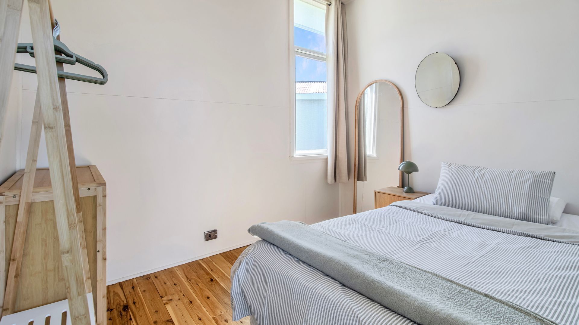 A bedroom with a wooden ladder, a bed with striped bedding, a bedside table with a lamp, a mirror, and a window with curtains.
