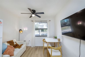 Ein Zimmer mit Deckenventilator, einem an der Wand befestigten Fernseher, einem Esstisch mit Stühlen, einer Couch mit Kissen und einem Fenster mit Vorhängen.