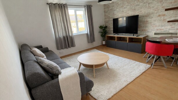 Ein modernes Wohnzimmer mit grauem Sofa, einem runden Couchtisch, einem Fernseher an einer Ziegelwand und einem Essbereich mit roten Stühlen.