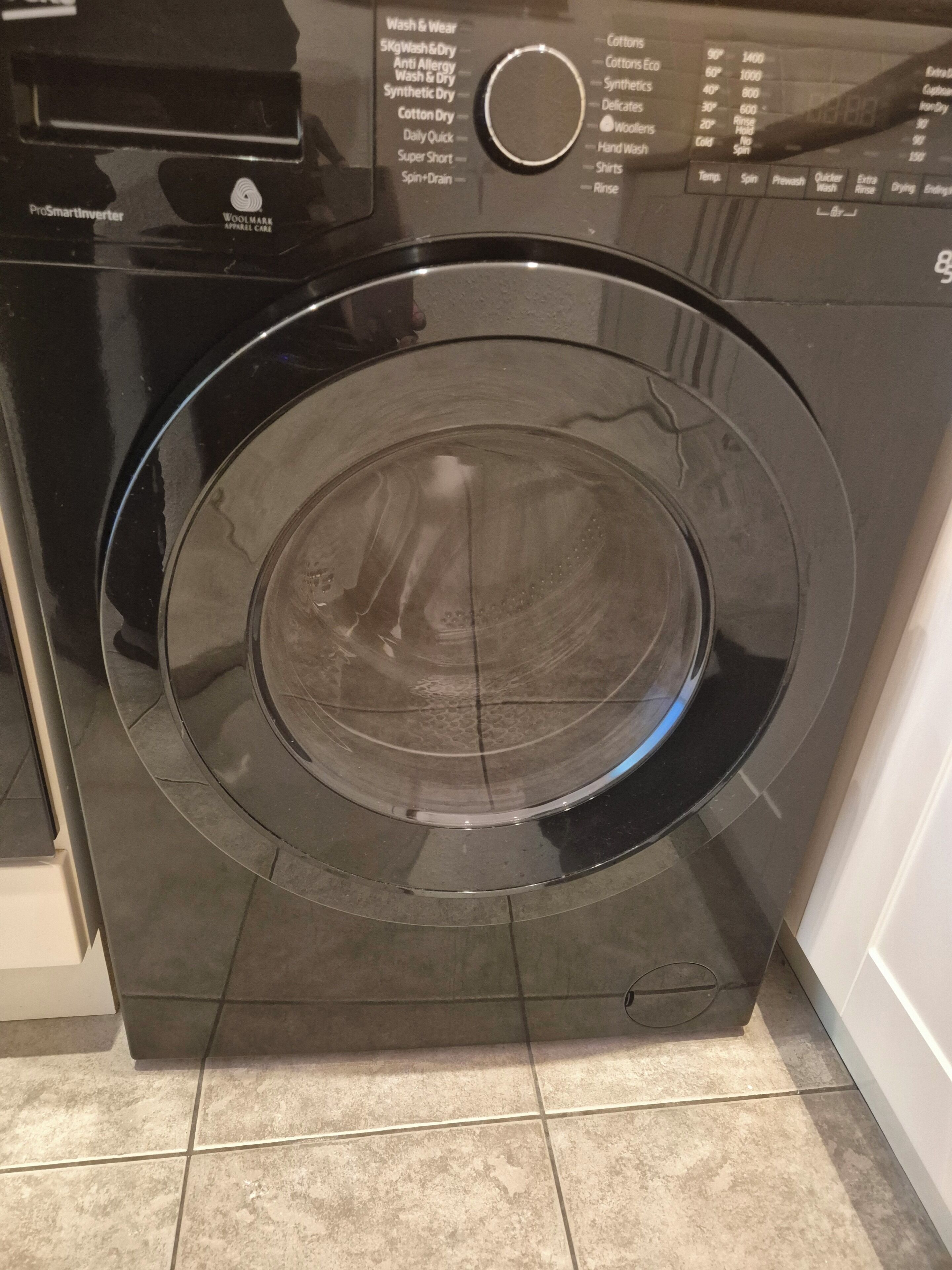 A front view of a black Woolmark branded washing machine with a control panel displaying various wash settings.