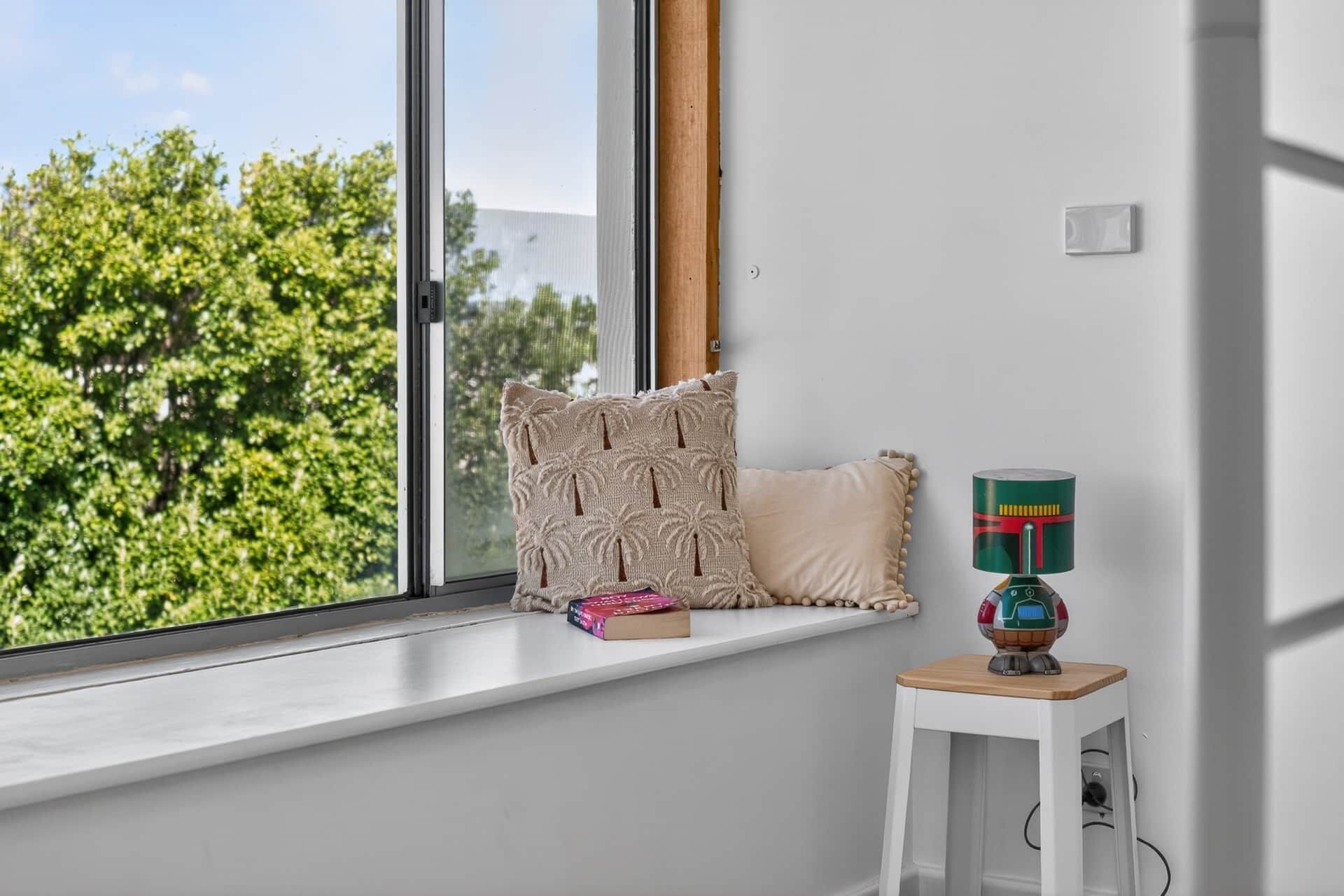 Een kamer met een groot raam, een witte wand, een klein wit tafeltje met een lamp en een kussen op de vensterbank.
