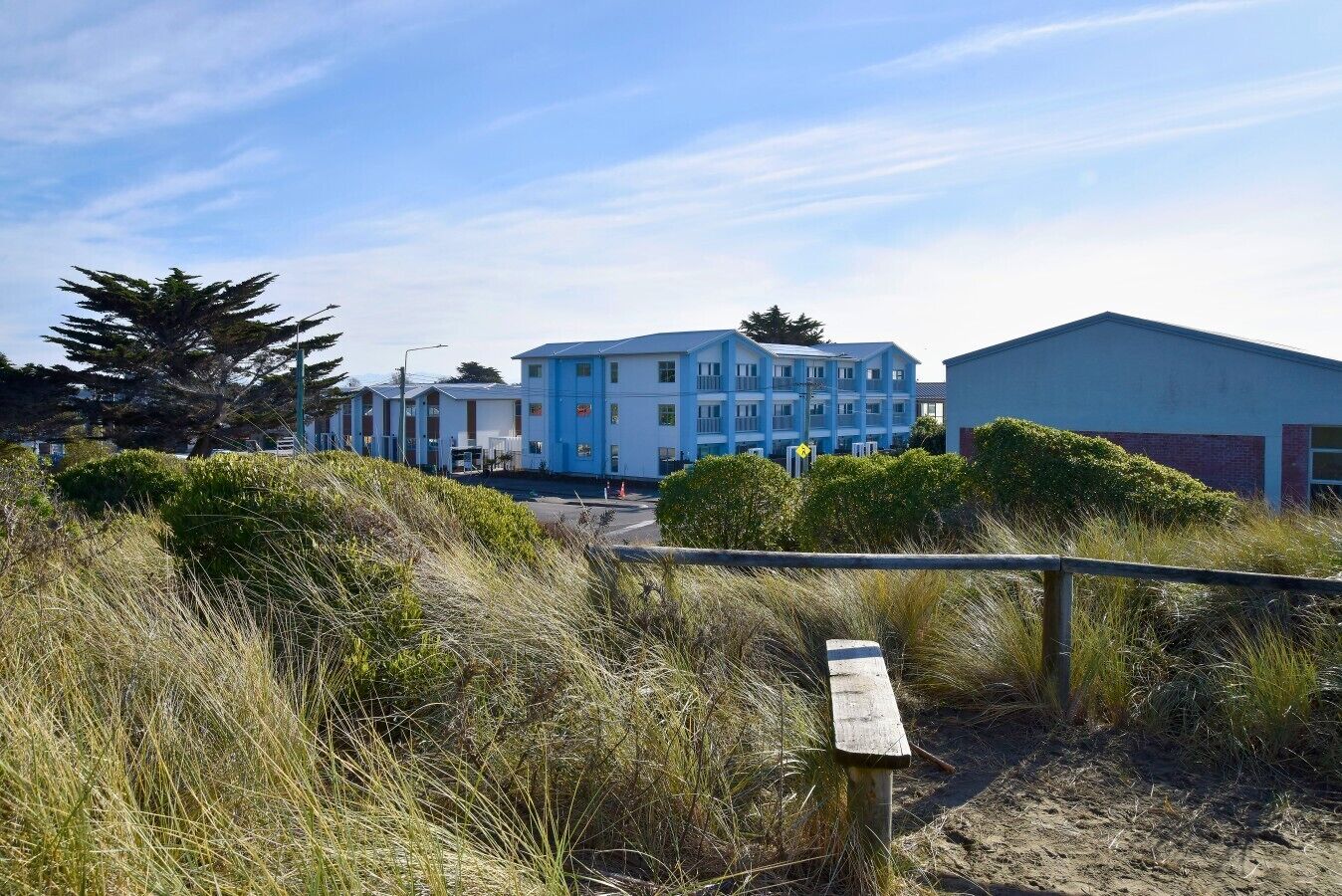 A coastal hotel with multiple blue buildings, a sandy beach, and a wooden walkway.